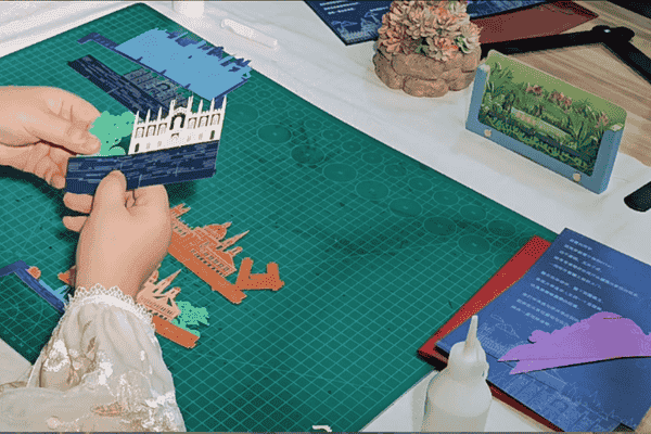 A photo of hands assembling a colorful pop-up paper art on a cutting mat with other designs in the background